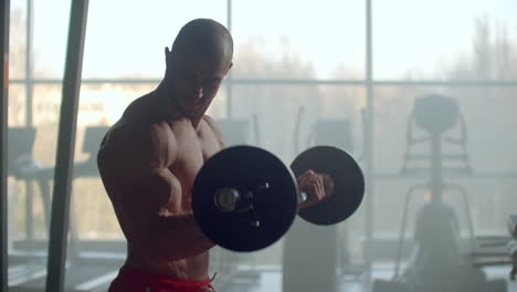 A-man-without-a-shirt-lifts-a-barbell-with-weight-performing-exercises-on-the-biceps.-Training-strong-hands-in-slow-motion-against-a-large-window-in-a-modern-stylish-fitness-center.-Bodybuilder-trains-hands.